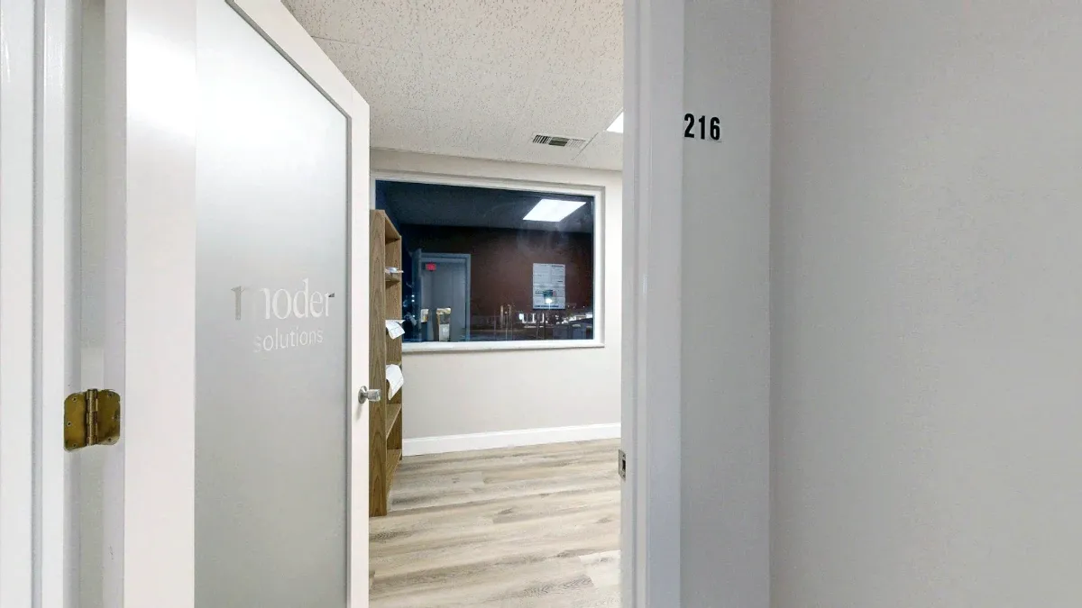 Open frosted-glass door to office 216 frames a large window, wooden shelving and light wood-look flooring.