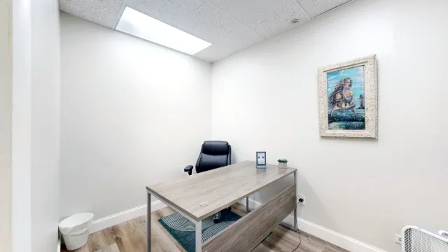 Office 217 viewed from the doorway, with a wood-look desk, black office chair, patterned rug and framed mermaid artwork.