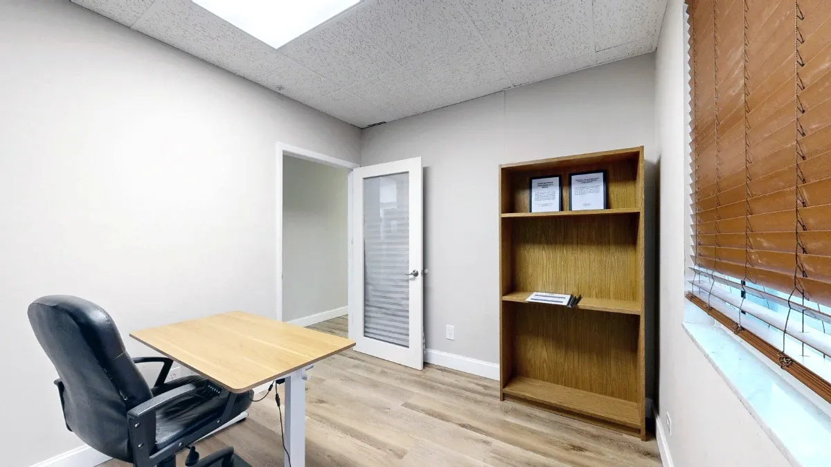Office 207 viewed toward the open door, with a tall bookshelf, desk and chair beside a window with brown blinds.