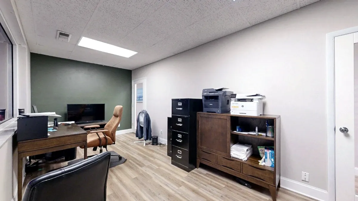 Office view facing a wooden storage cabinet with printers, black filing cabinets, two doors, and a desk along the windows.