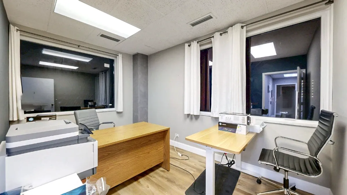 Office 223A interior with curtained windows on two walls, a wooden desk, a smaller desk, two chairs and a printer.