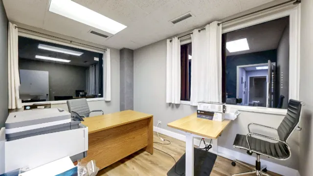 Office 223A interior with curtained windows on two walls, a wooden desk, a smaller desk, two chairs and a printer.