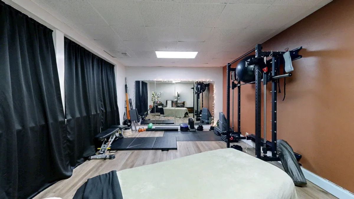 Office 227 exercise area with a tall black rack, bench, floor mats and a wall mirror beyond the massage table.