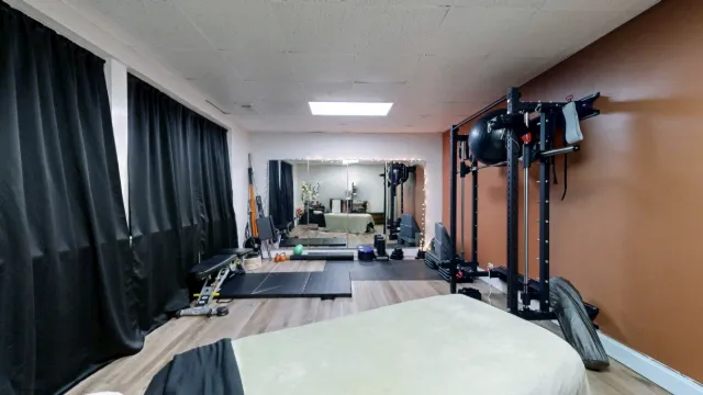 Office 227 exercise area with a tall black rack, bench, floor mats and a wall mirror beyond the massage table.