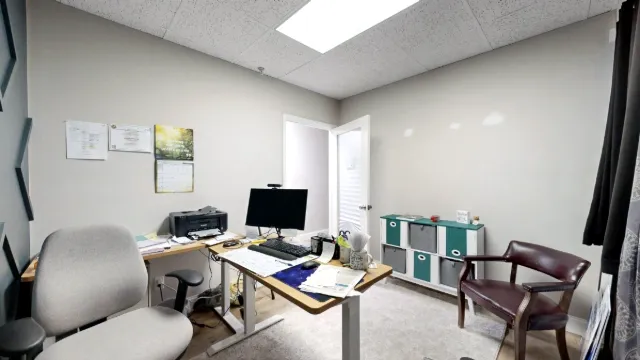 Wider view of Office 211 showing two chairs, a paper-covered desk, a light rug, and dark curtains along the right edge.