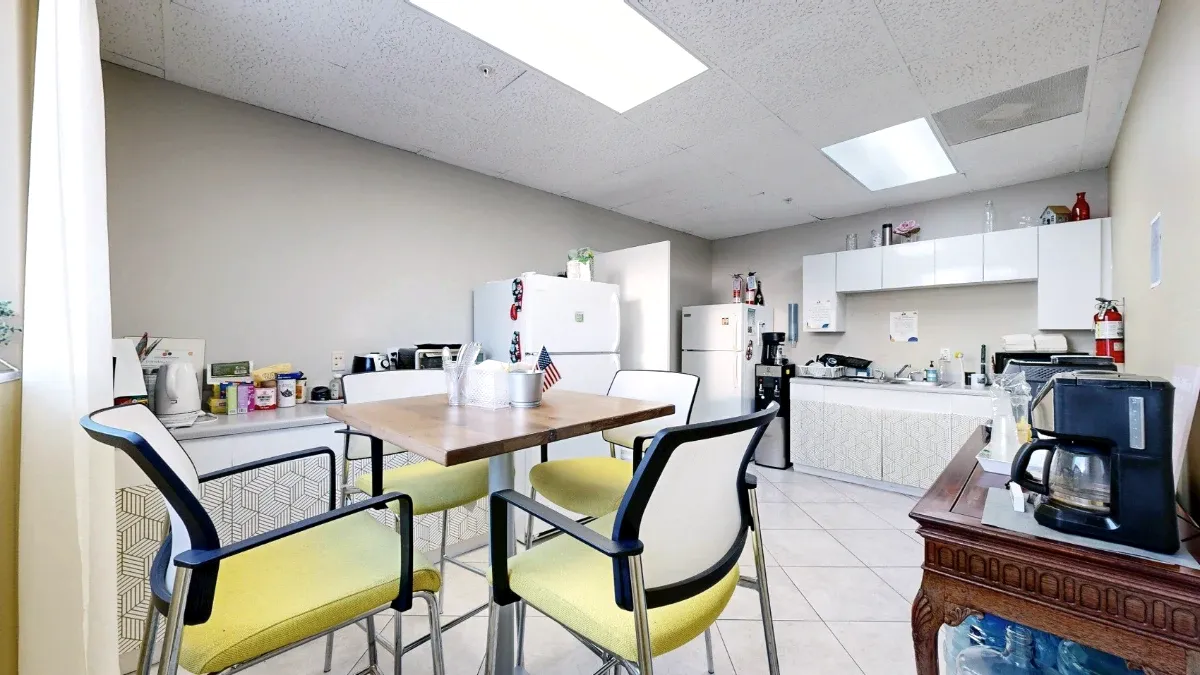 Yellow-cushioned chairs surround a lunch table, with two refrigerators, sink cabinetry and a foreground coffee maker.
