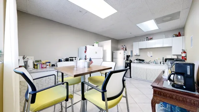 Yellow-cushioned chairs surround a lunch table, with two refrigerators, sink cabinetry and a foreground coffee maker.