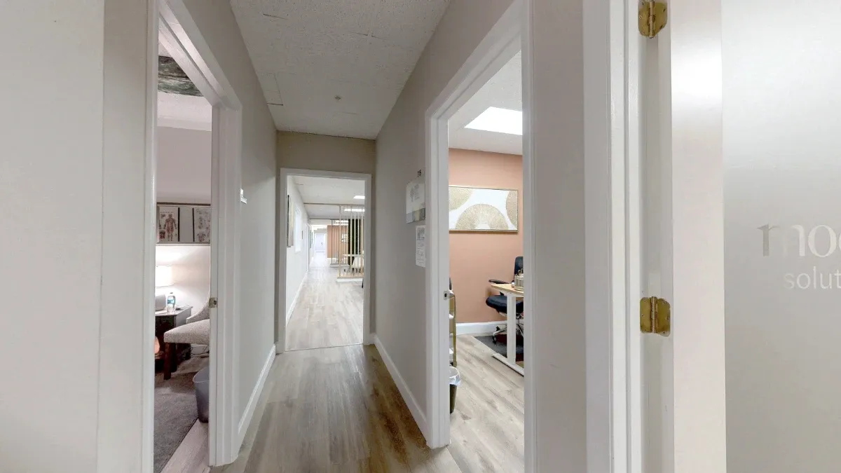 Hallway approach to Office 215 with a partial view of its desk, black chair and peach accent wall through the right doorway.