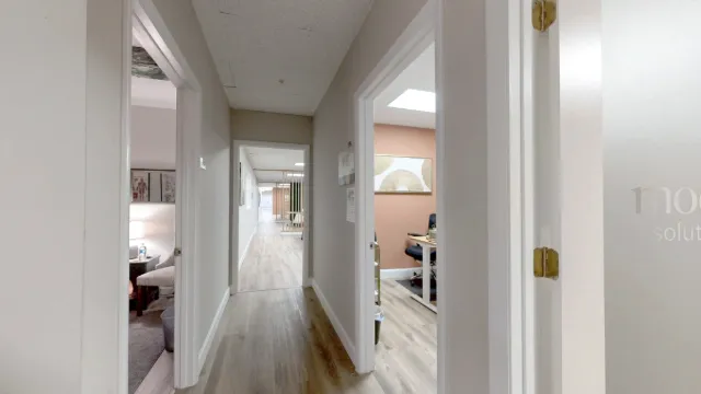 Hallway approach to Office 215 with a partial view of its desk, black chair and peach accent wall through the right doorway.