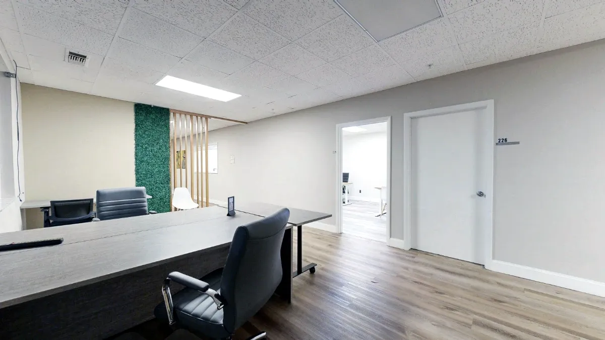 Shared desks and black office chairs in an open coworking area with wood-look flooring and a green accent divider.