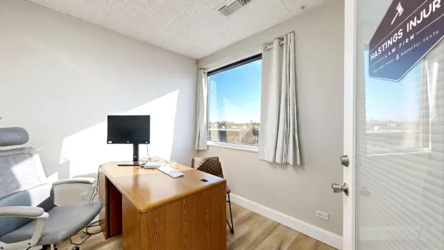 Small office with a wooden desk, monitor, gray task chair, guest chair, curtained window and glass door.