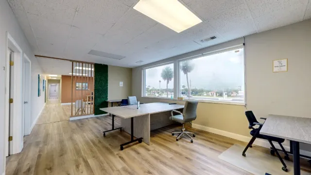 Grouped gray desks and rolling chairs sit below two windows, beside a corridor and a wood-slat divider.