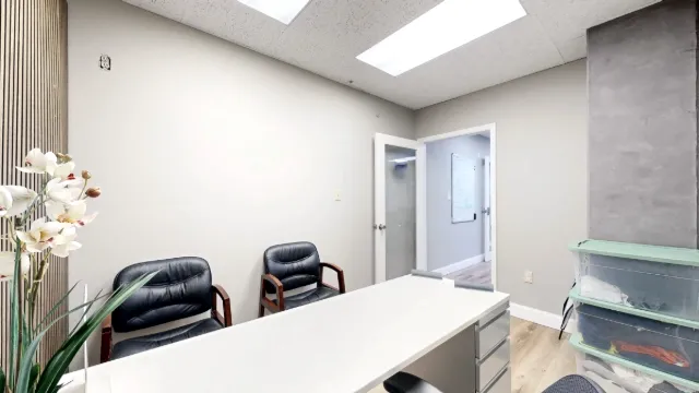 View across a white desk toward two black guest chairs and an open door, with orchids at left and storage bins at right.