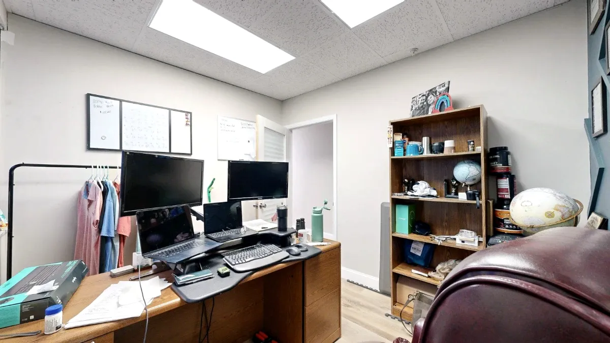 Reverse view of Office 212 showing desk monitors and laptops, a bookshelf, whiteboards and a clothing rack near the door.