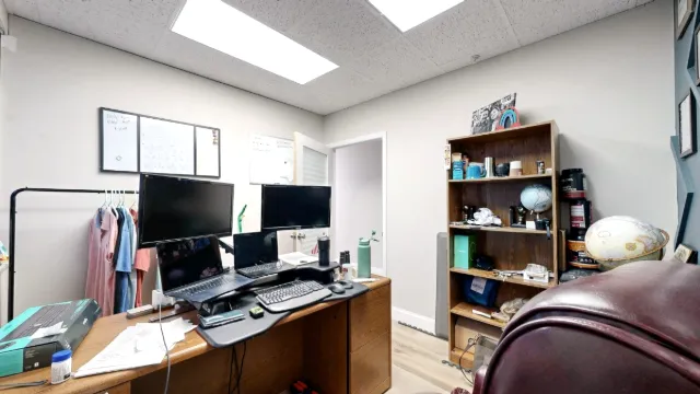 Reverse view of Office 212 showing desk monitors and laptops, a bookshelf, whiteboards and a clothing rack near the door.