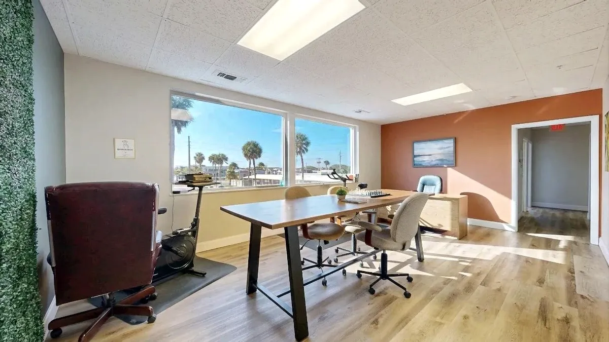 A chessboard rests on a shared table as sunlight crosses beige chairs and the floor beside an orange accent wall.