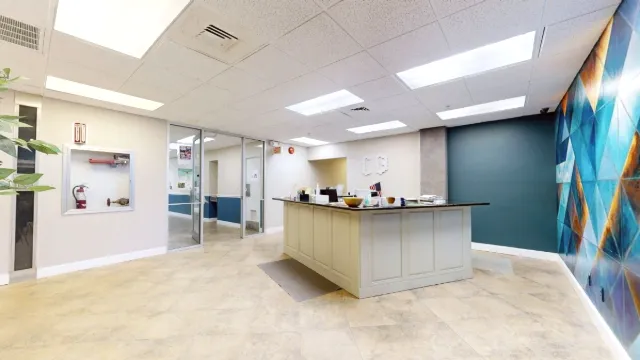 Reception desk viewed diagonally toward the glass entrance, with a fire equipment cabinet on the left and mural on the right.