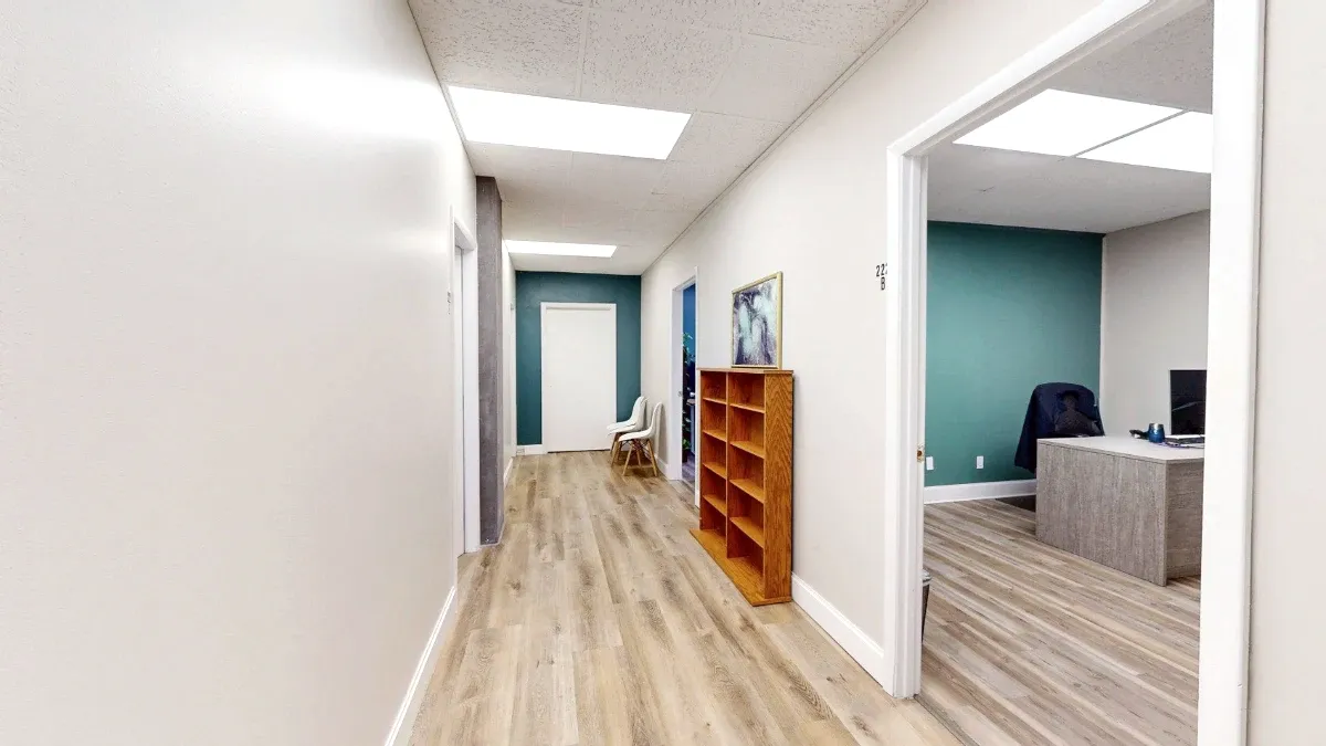 Interior hallway with wooden cubby shelving, white chairs at the far end, and an open office doorway on the right.