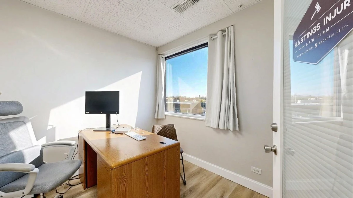 Sunlit office with a wooden desk, monitor, ergonomic chair, guest chair, and a large curtained window.