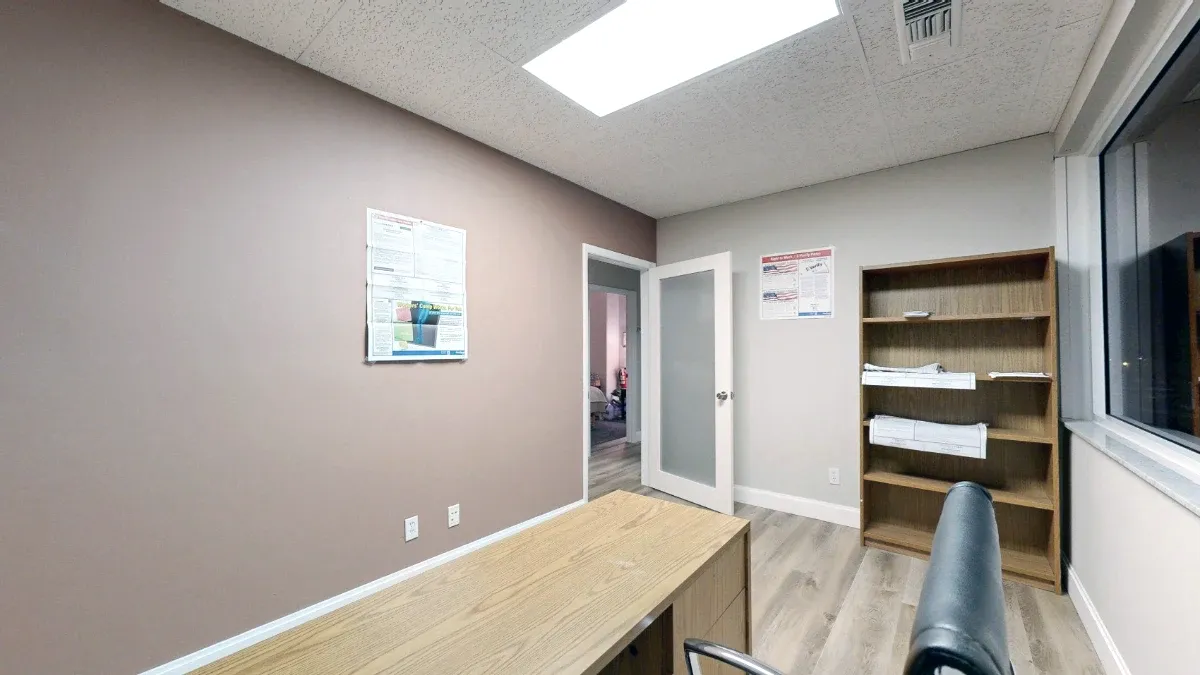View past the desk and black chair toward office 216's open door, tall wooden bookcase and posted wall notices.