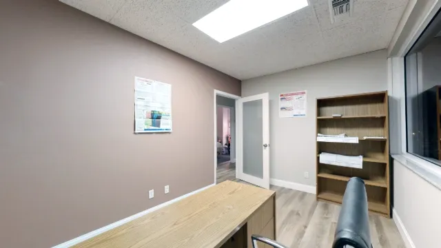 View past the desk and black chair toward office 216's open door, tall wooden bookcase and posted wall notices.
