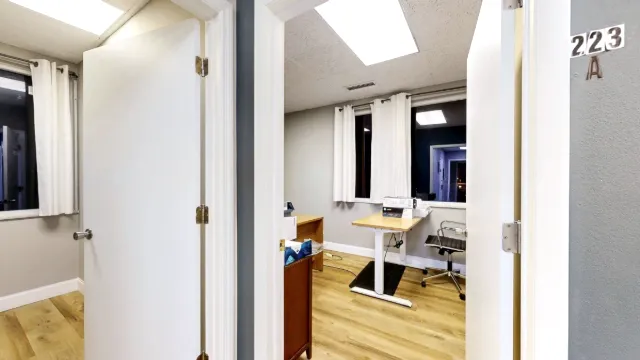View through the open doorway of Office 223A, with wall signage, a small desk, a rolling chair and white window curtains.