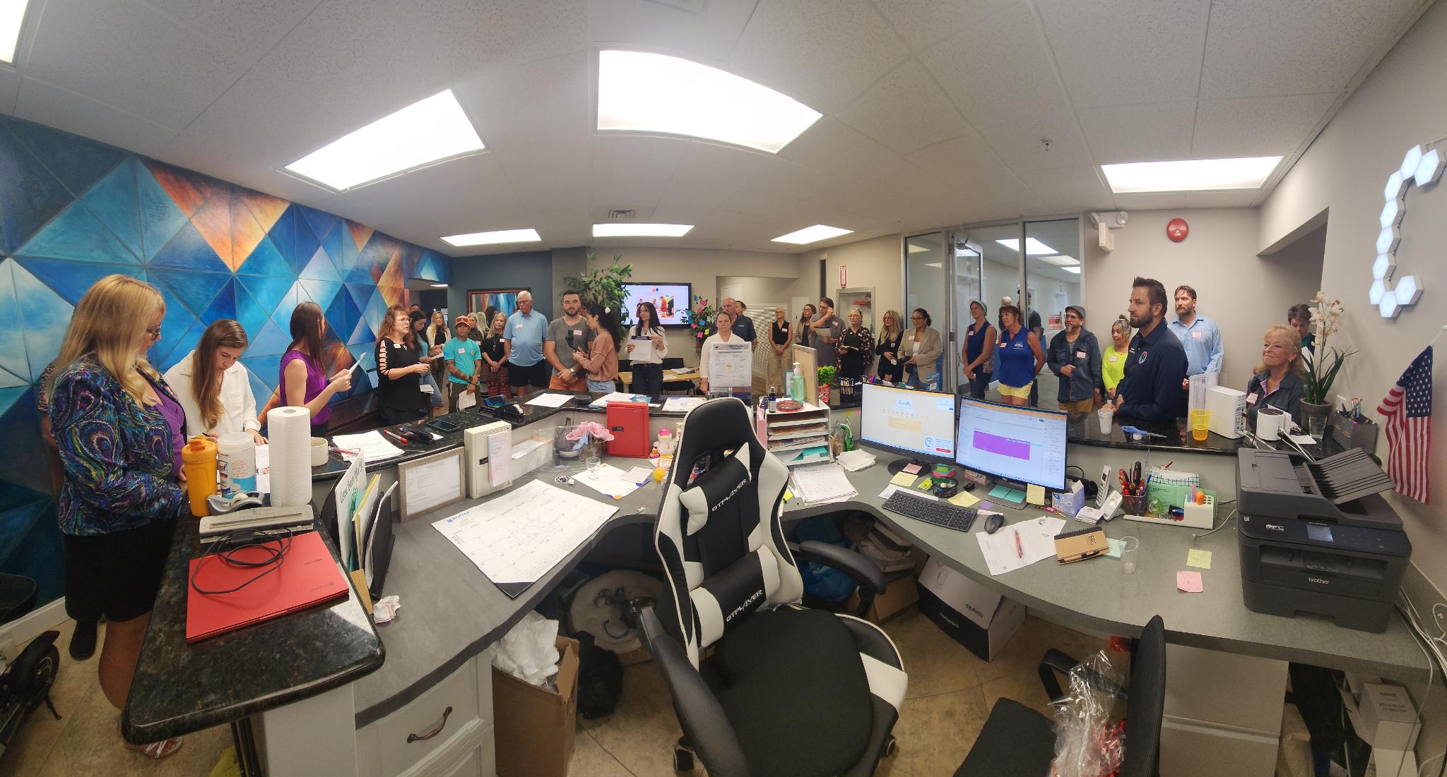 A large group wearing name tags gathers around a reception desk beside a blue geometric mural.