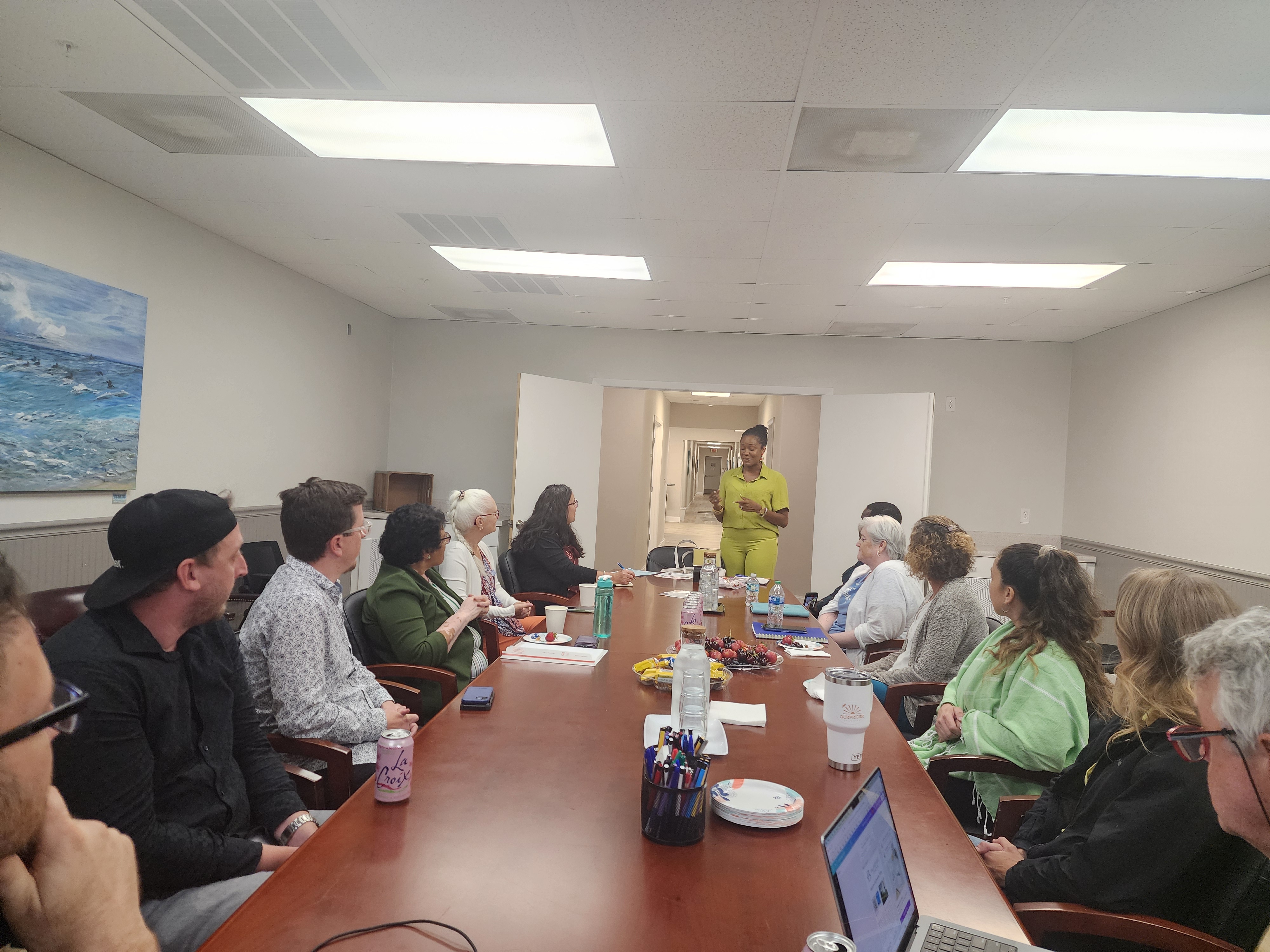 A standing speaker in green addresses attendees seated around a long conference table with refreshments and notebooks.
