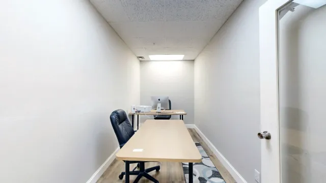 A straight view down the narrow office shows a long desktop, black chair and computer desk at the far wall.