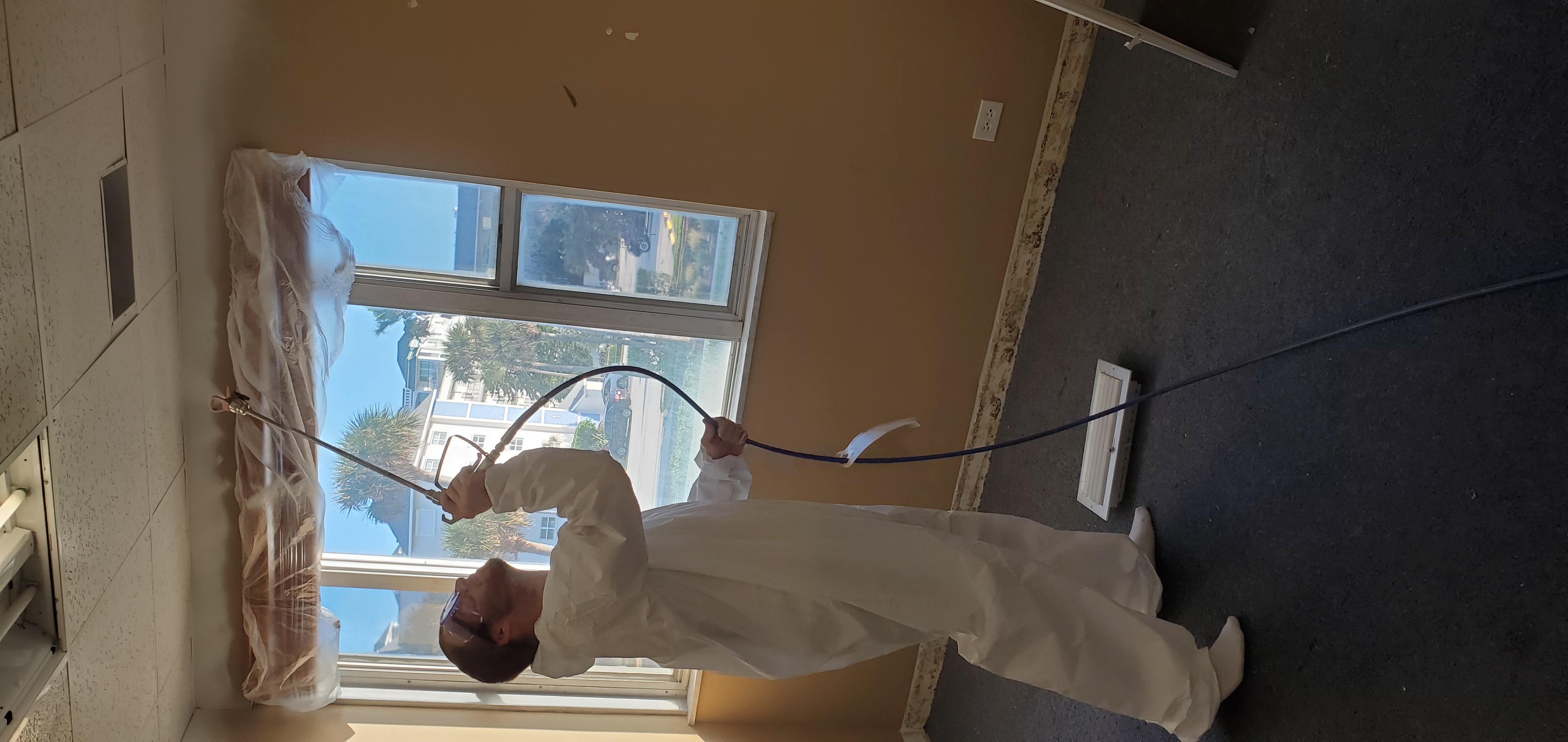 Robbie wears white painting coveralls and goggles while aiming a paint sprayer at ceiling tiles beside a large window.
