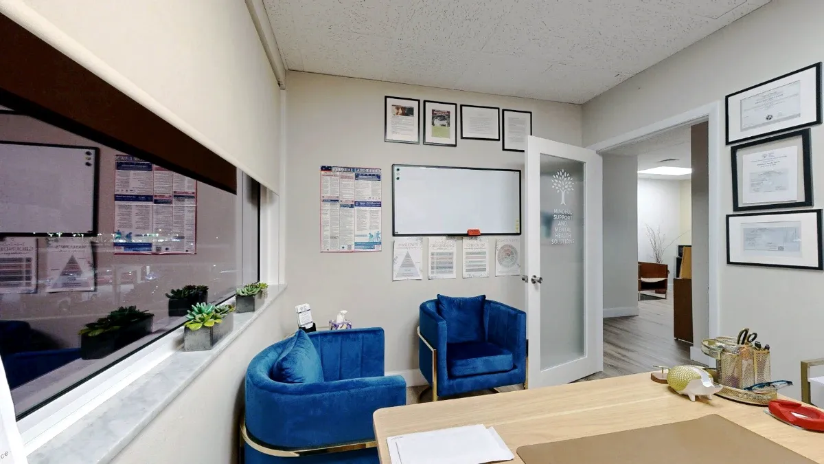 View across the desk toward two blue armchairs, a window, a whiteboard and the open frosted-glass door in Office 215.