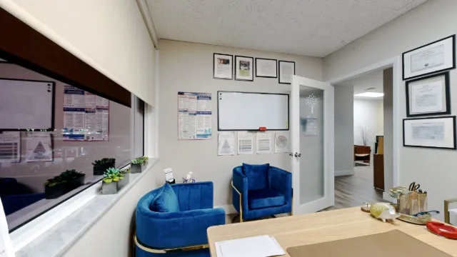 View across the desk toward two blue armchairs, a window, a whiteboard and the open frosted-glass door in Office 215.