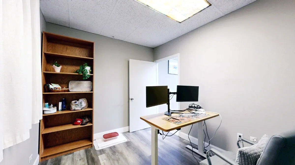 Reverse view of the office showing two monitor screens, tall wooden shelving, an open door and a gray chair.