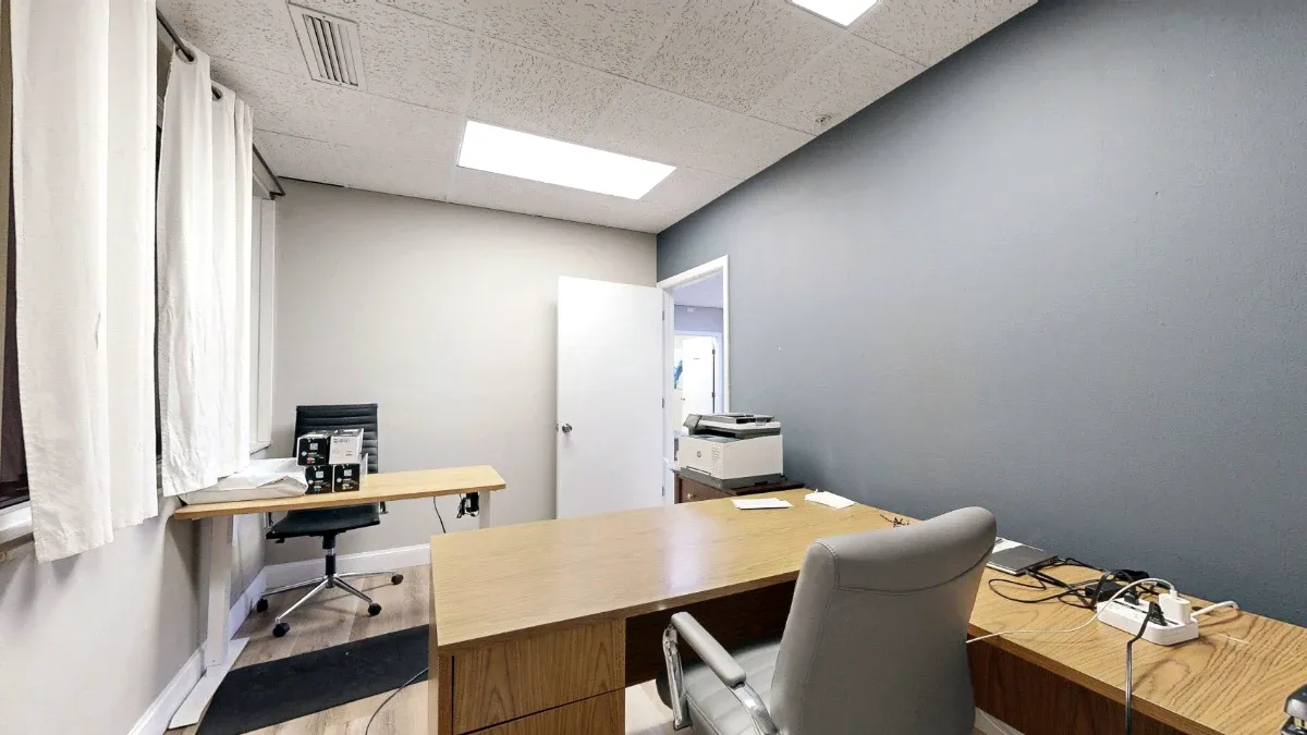 Office with wood desks, two chairs, white-curtained windows, a printer and a gray accent wall.