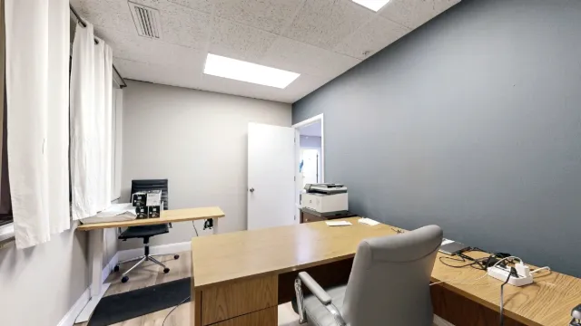 Office with wood desks, two chairs, white-curtained windows, a printer and a gray accent wall.