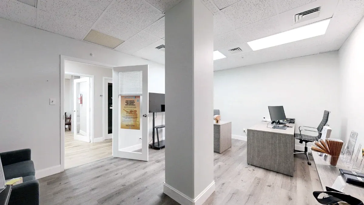Office 226 interior with a gray desk, monitor and black chair to the right of a central column, facing an open entrance.