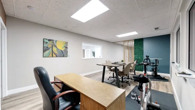 A wooden desk fills the foreground, with two exercise bikes along the windows and a shared table near a teal wall.