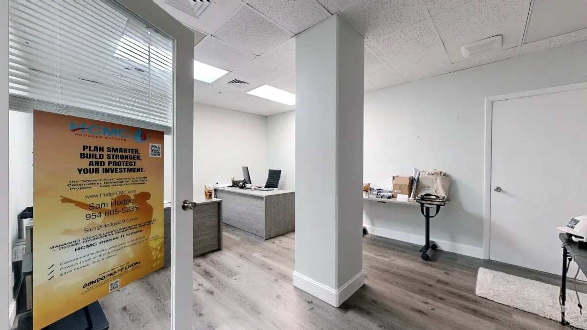 View through Office 226's open glass door toward a central column, gray desks, a side worktable and a closed white door.