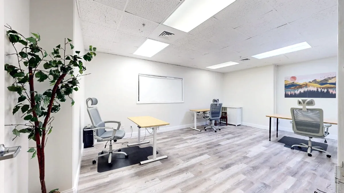 Wide doorway view of Office 229 with three desks, gray chairs, a shaded interior window and landscape artwork.