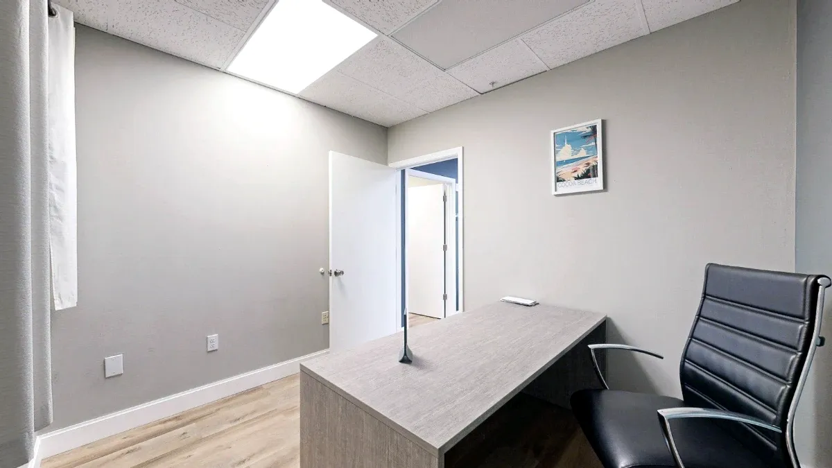 Compact office with a wood-look desk, black chair, curtained window, open door and framed Cocoa Beach print.
