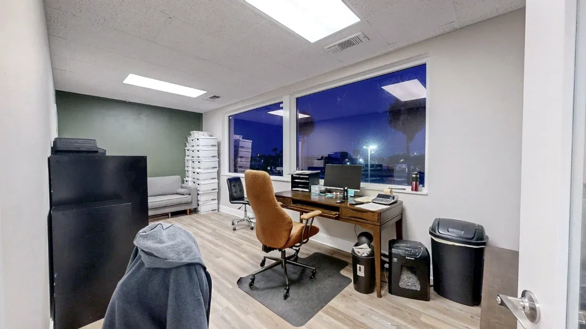 Doorway view of an office with two windows, a wooden desk, a tan swivel chair, and a gray sofa at the back.