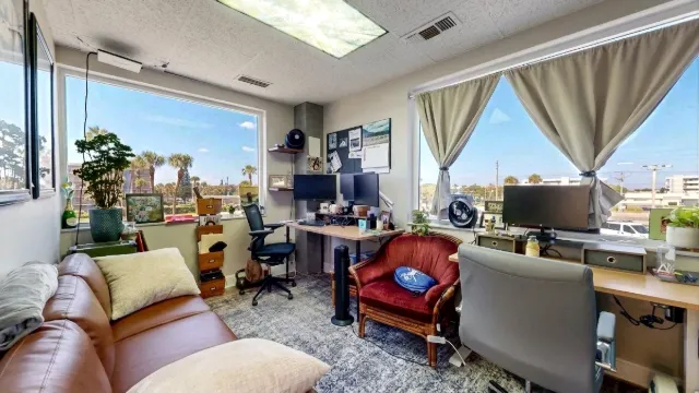 Window-side desks with monitors surround a red armchair, while a brown sofa and patterned rug fill the foreground.