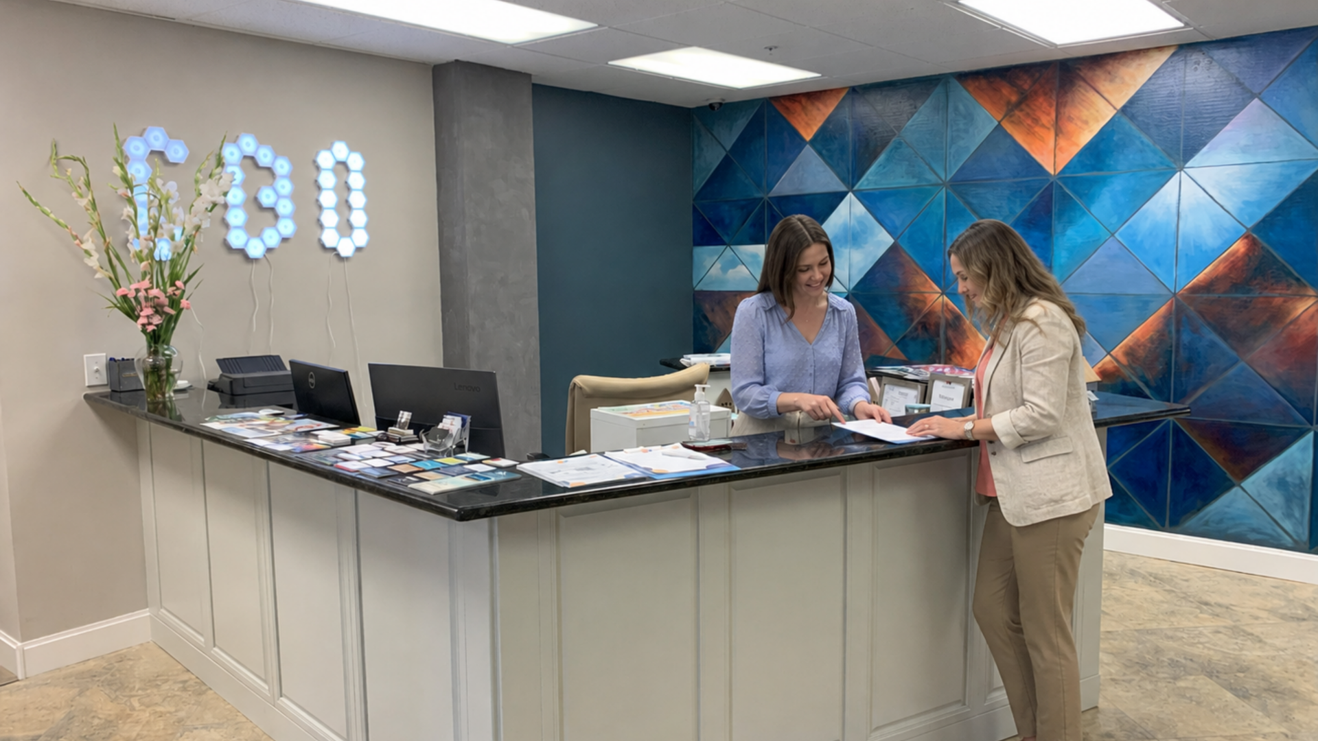 Two women review paperwork at a reception desk with illuminated CBO letters and a blue geometric mural behind them.