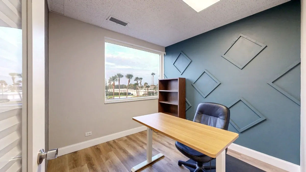 Sunlit office with a wood desk, black chair, bookshelf, blue geometric accent wall, and a window overlooking palm trees.