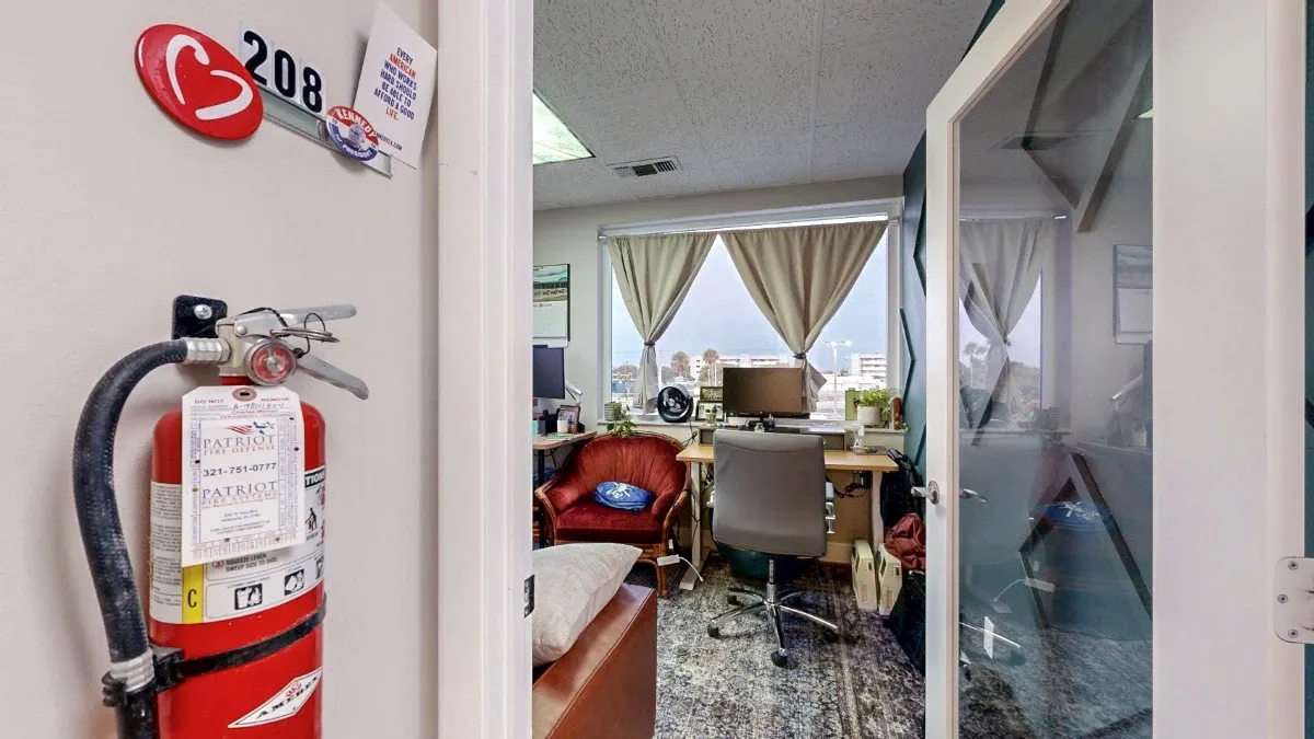 Open glass door to Office 208 reveals a window-facing desk, gray swivel chair, red accent chair and patterned rug.