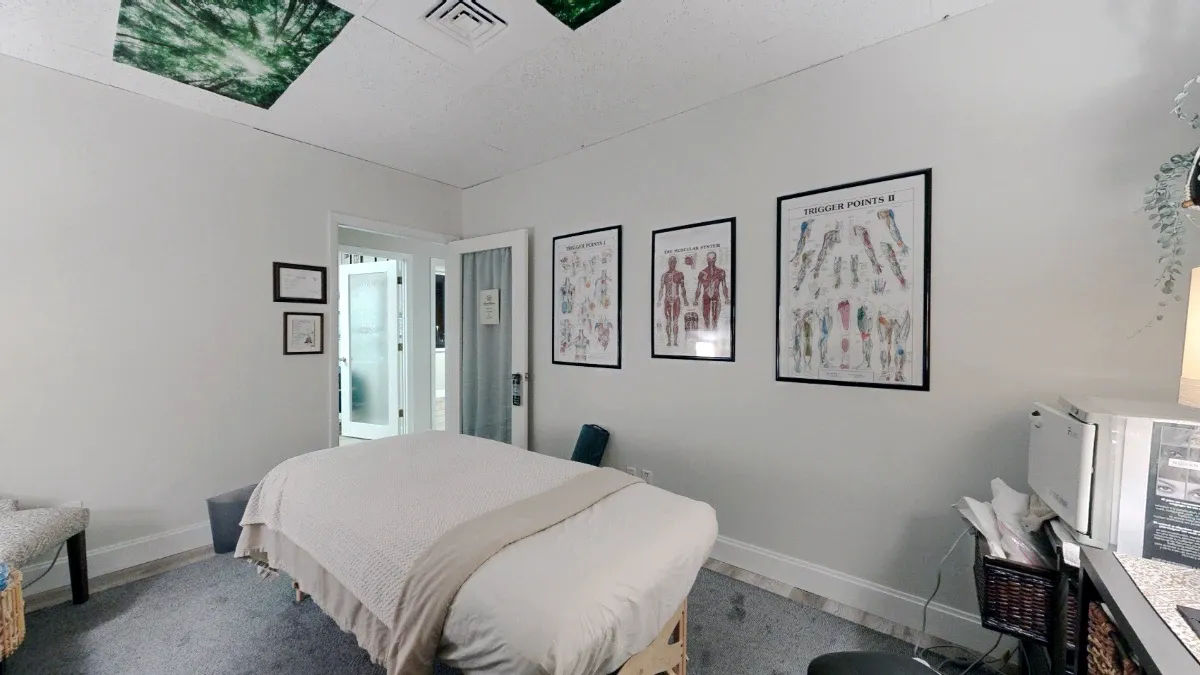 Reverse view across the massage table toward an open door, with three framed anatomy charts on the wall and storage at right.