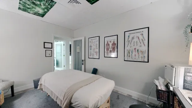 Reverse view across the massage table toward an open door, with three framed anatomy charts on the wall and storage at right.