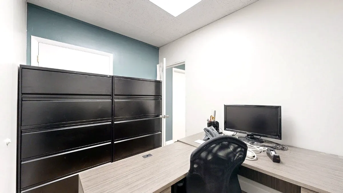 Office with an L-shaped desk, mesh chair, monitor, phone and tall black filing cabinets against a blue-gray wall.