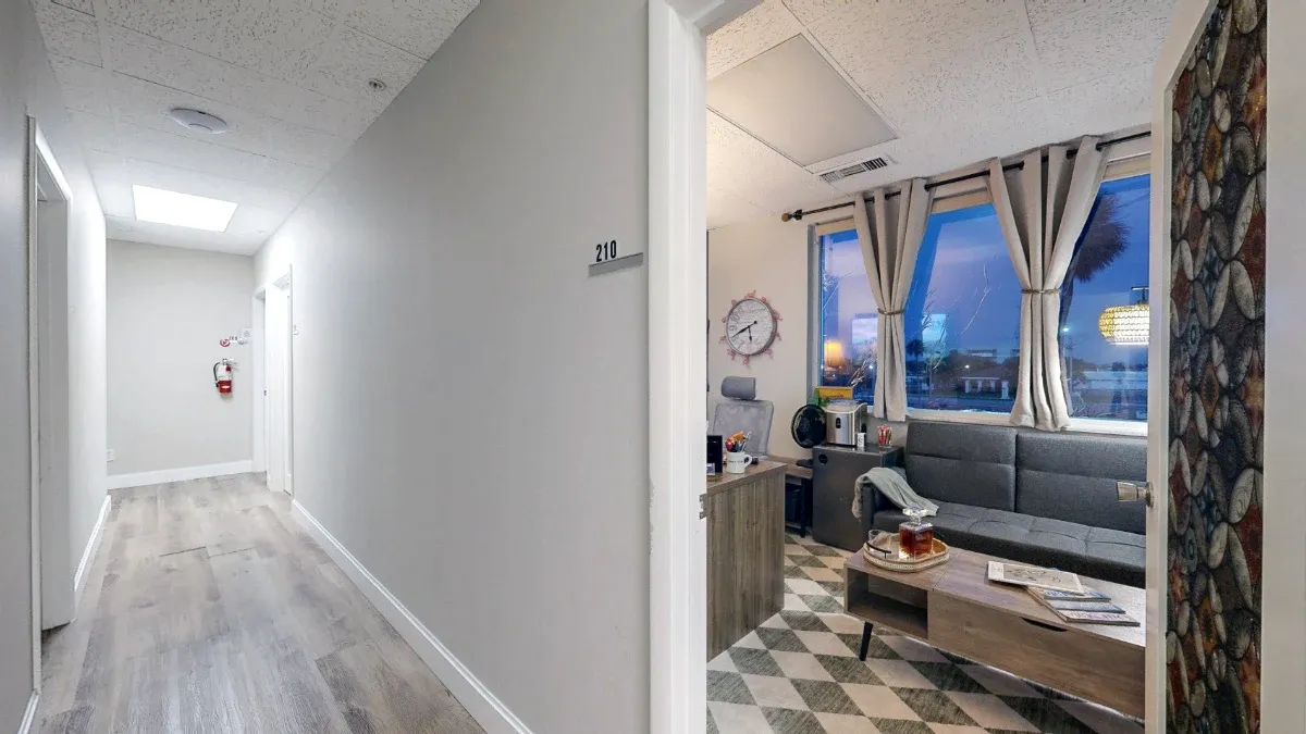 Hallway view through the open office door shows a gray sofa, coffee table, curtained windows and part of the desk.