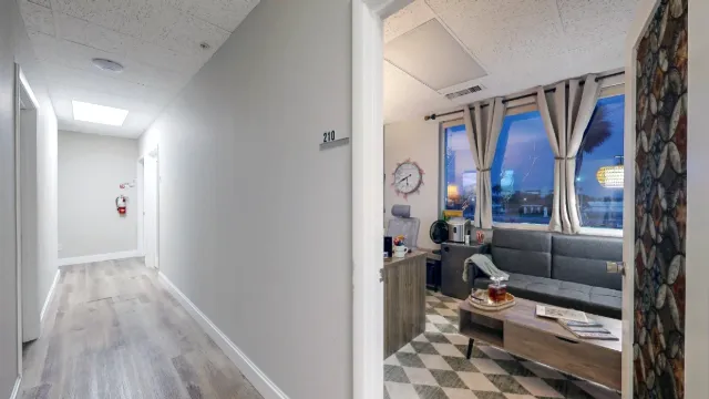 Hallway view through the open office door shows a gray sofa, coffee table, curtained windows and part of the desk.